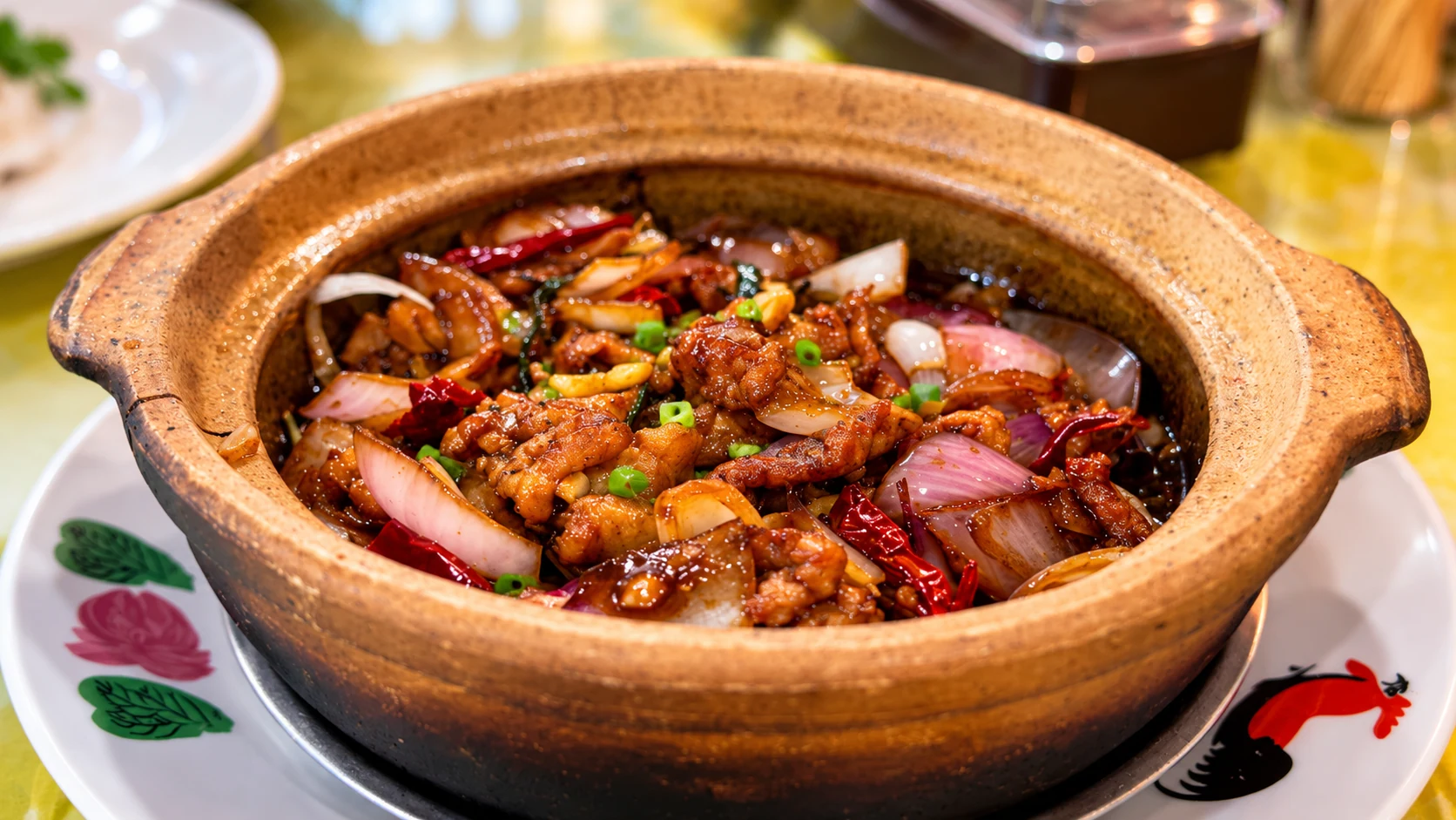 A brown claypot with glossy cooked pieces, onion and red strips.